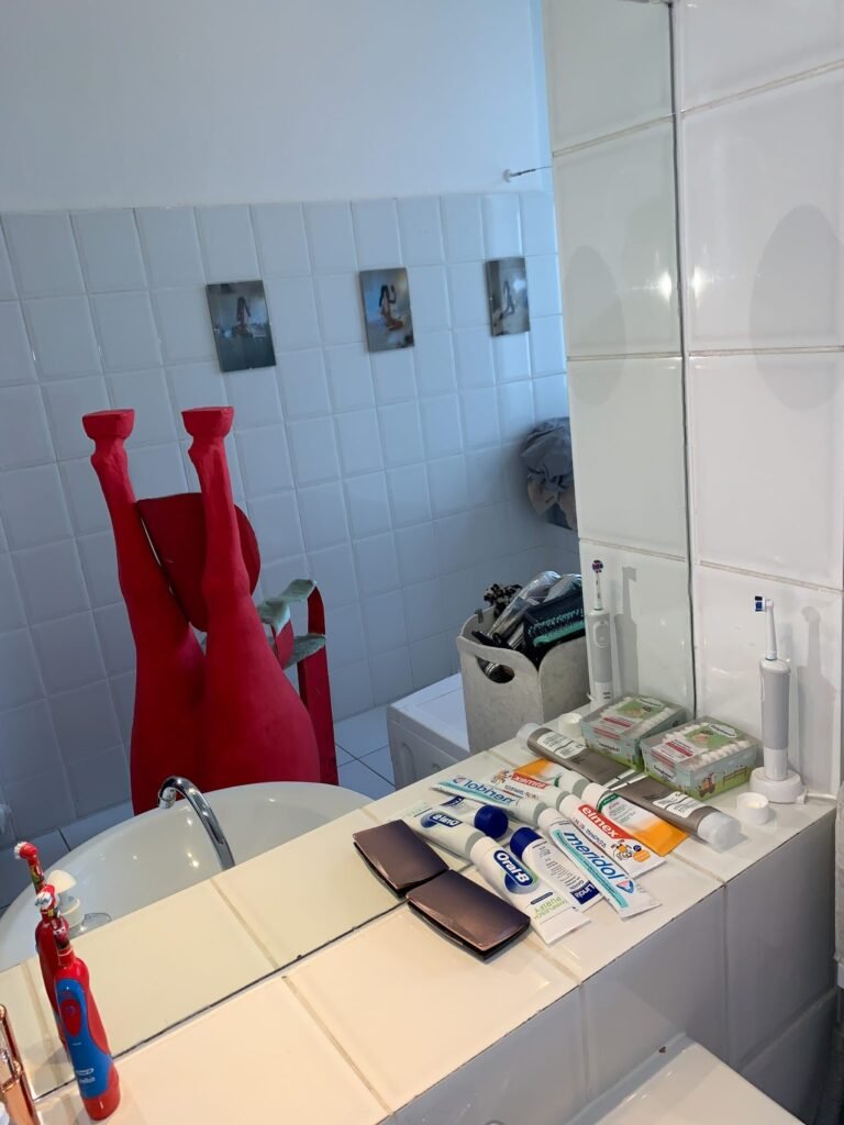 Escultura roja abstracta en baño moderno blanco con lavabo y productos de higiene, obra de Horacio Alcolea Crespo, arte contemporáneo.
